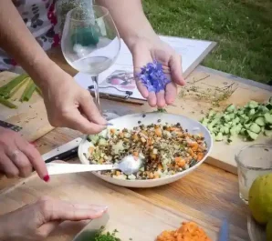 Des mains ajoutent des fleurs de bourrache bleues à une salade colorée de lentilles, concombre et carotte. Scène d'un atelier Les Vivantes où l'on apprend à préparer des plats d'alimentation vivante avec des ingrédients frais et bio, y compris des fleurs comestibles.